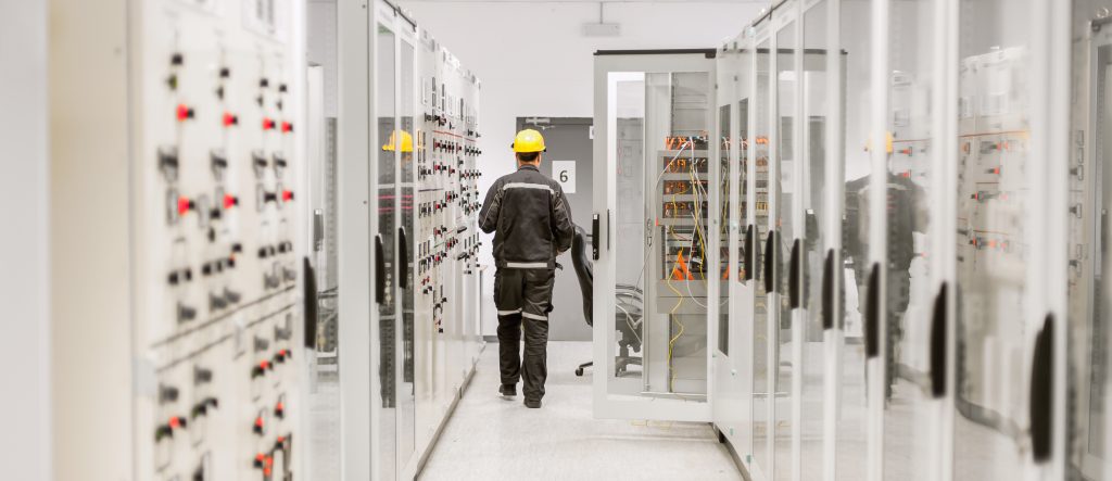 Man in PPE walking through a clean, modern, control centre for an electricity/energy plan. Copyright image used under Dreamstime license to Darrell Morrison Graphic Designn.